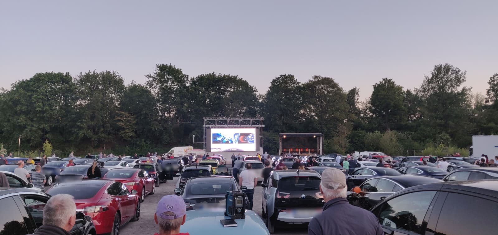 Reihen geparkter Elektroautos in der Abenddämmerung vor einer großen LED-Leinwand auf einem Schotterplatz