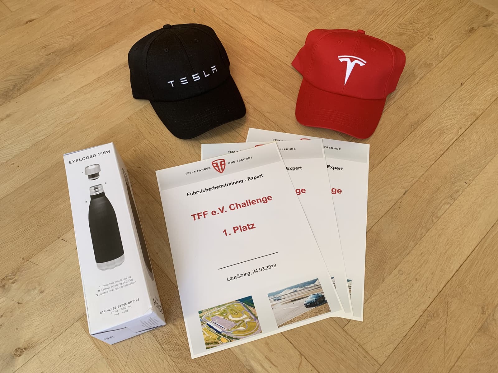 Mehrere Urkunden der TFF-Challenge Lausitzring 2019, daneben zwei Tesla-Kappen und eine Thermosflasche