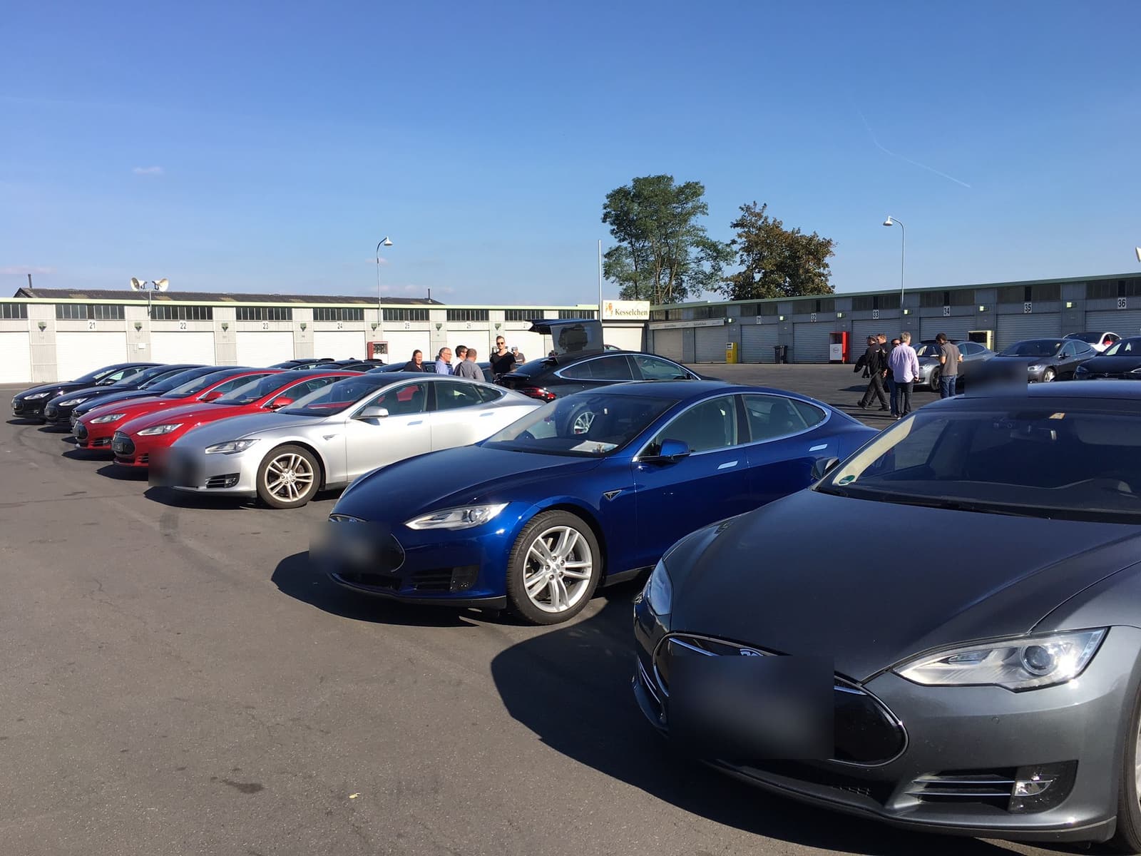 Lange Reihe geparkter Teslas im Fahrerlager des Nürburgrings, dahinter die Boxengaragen bei blauem Himmel