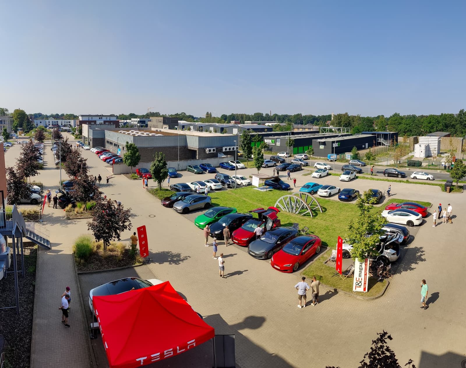 Blick von oben auf das Veranstaltungsgelände mit vielen geparkten Teslas, Besuchern und einem roten Zelt