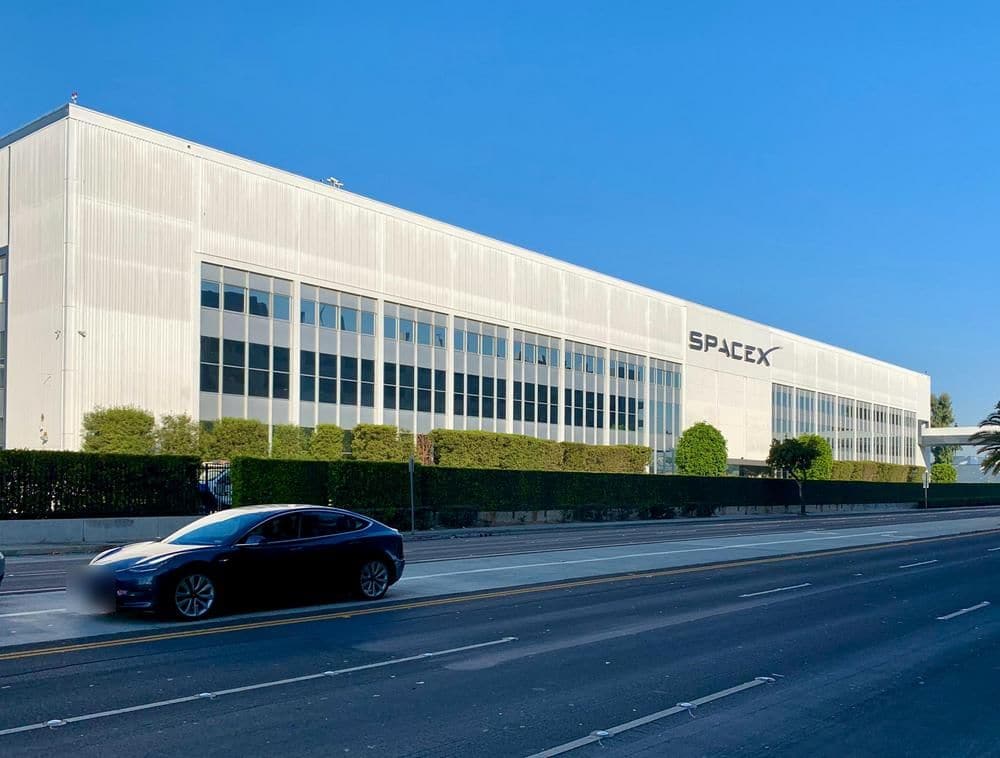 Langgestrecktes Firmengebäude von SpaceX mit Schriftzug an der Fassade, davor fährt eine dunkle Tesla-Limousine vorbei