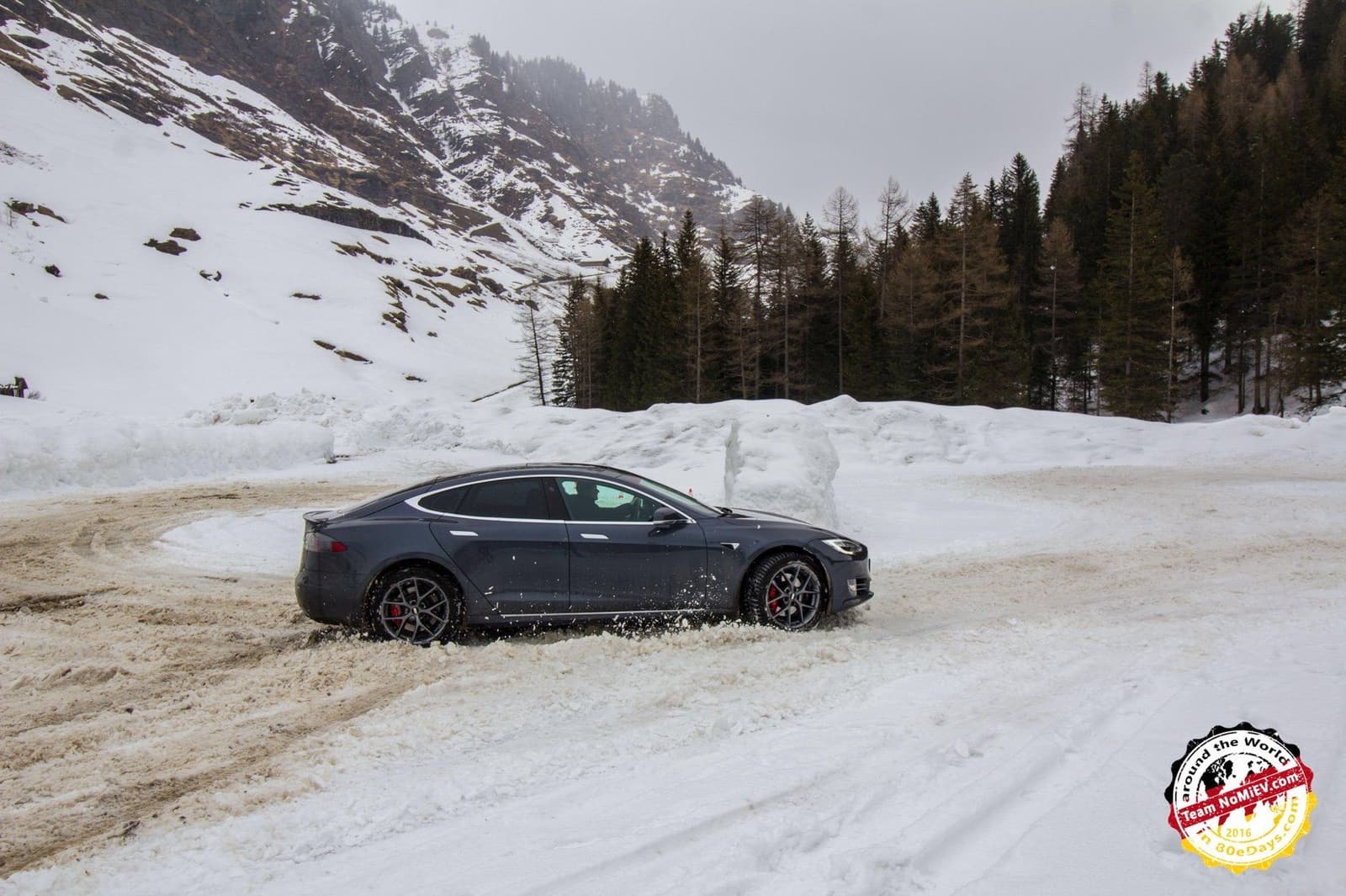 Dunkler Tesla Model S driftet über die verschneite Übungsfläche, dahinter Bergflanke und Nadelwald