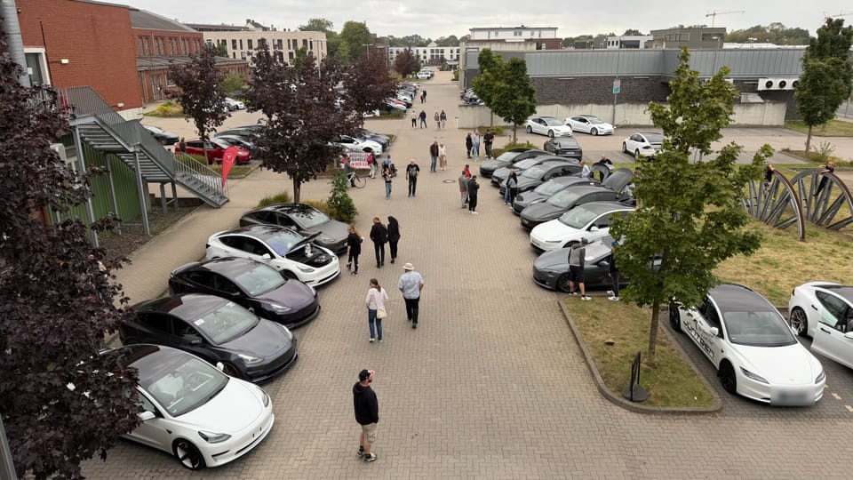 Reihe geparkter Teslas von oben, daneben ein blühender Baum und das Backsteingebäude