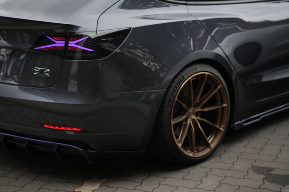 Heckpartie eines dunklen Tesla mit violett beleuchteten Rückleuchten und Bronzefelgen