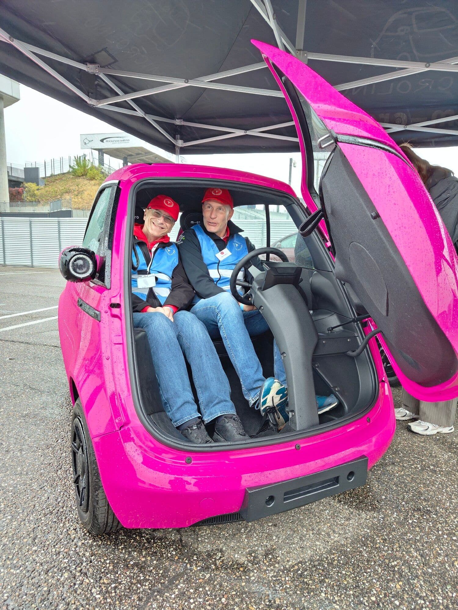 Zwei Helfer des Vereins in blauen Warnwesten sitzen nebeneinander im pinkfarbenen Microlino, die Fronttür steht offen
