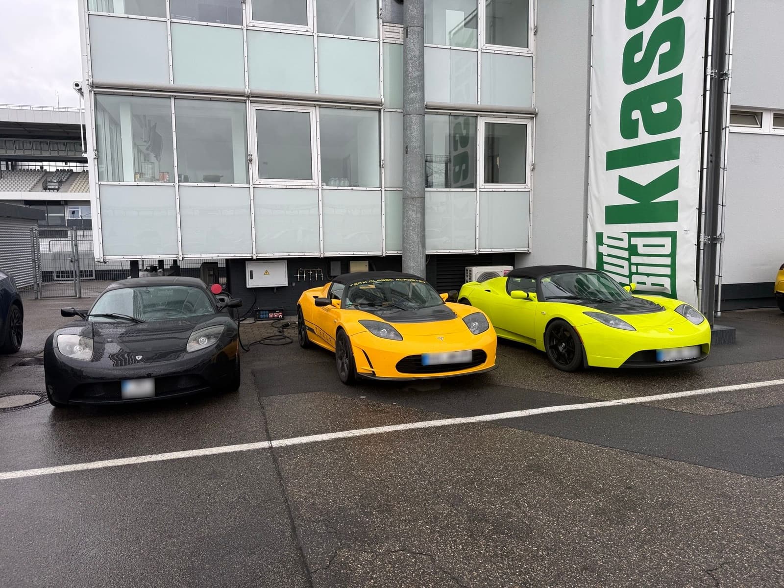 Drei Tesla Roadster in Schwarz, Gelb und Hellgrün nebeneinander vor dem Gebäude an der Boxengasse