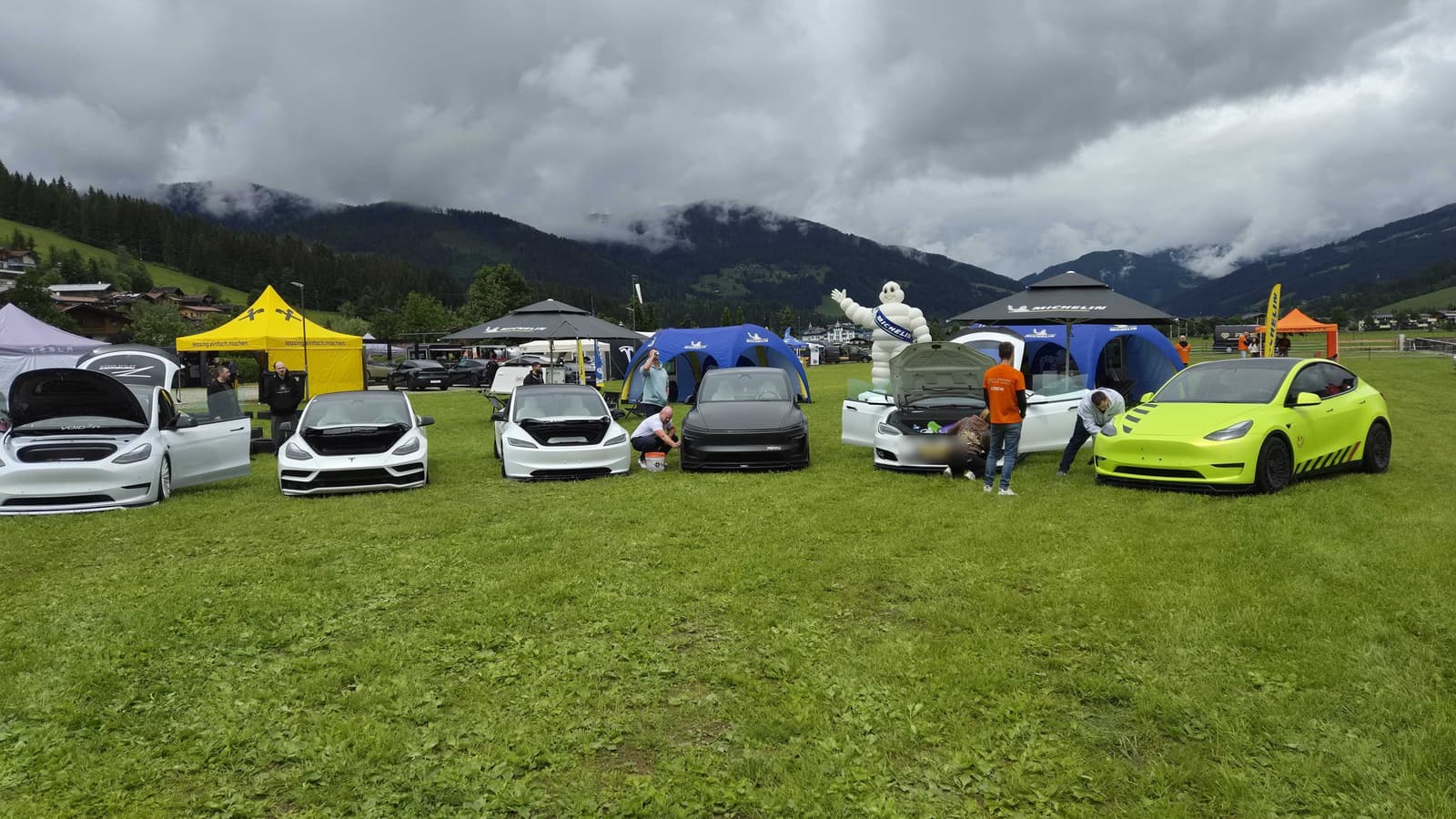 Reihe umgebauter Tesla-Fahrzeuge auf einer Wiese vor den Zelten der Aussteller, ganz rechts ein grellgrüner Model Y, dahinter die Berge