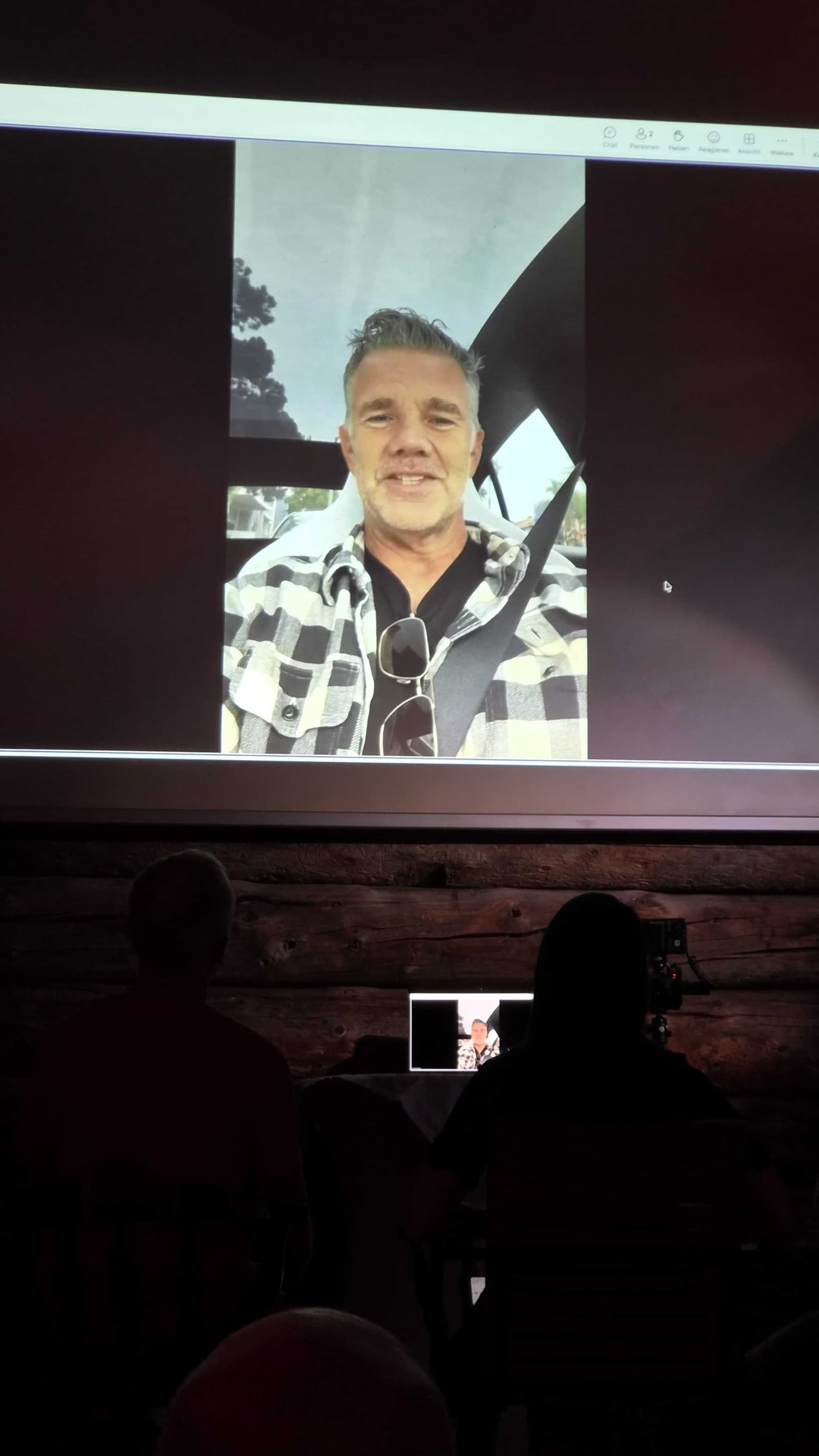 Leinwand im abgedunkelten Saal, darauf ein Teilnehmer der Live-Schaltung, davor die Silhouetten des Publikums