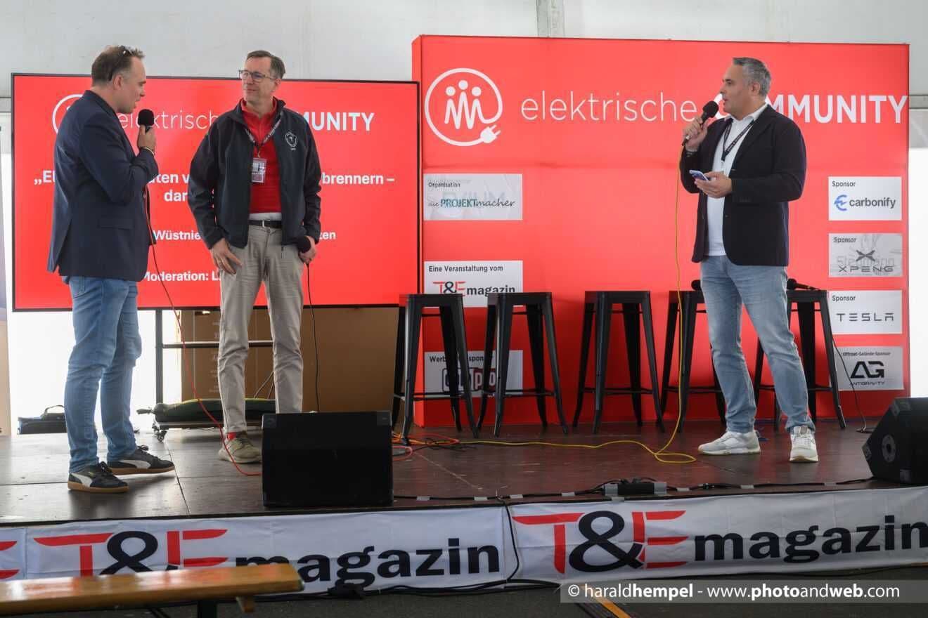 Drei Männer mit Mikrofonen auf der Bühne der Elektrischen Community, in der Mitte ein Vertreter des Vereins in der Vereinsjacke
