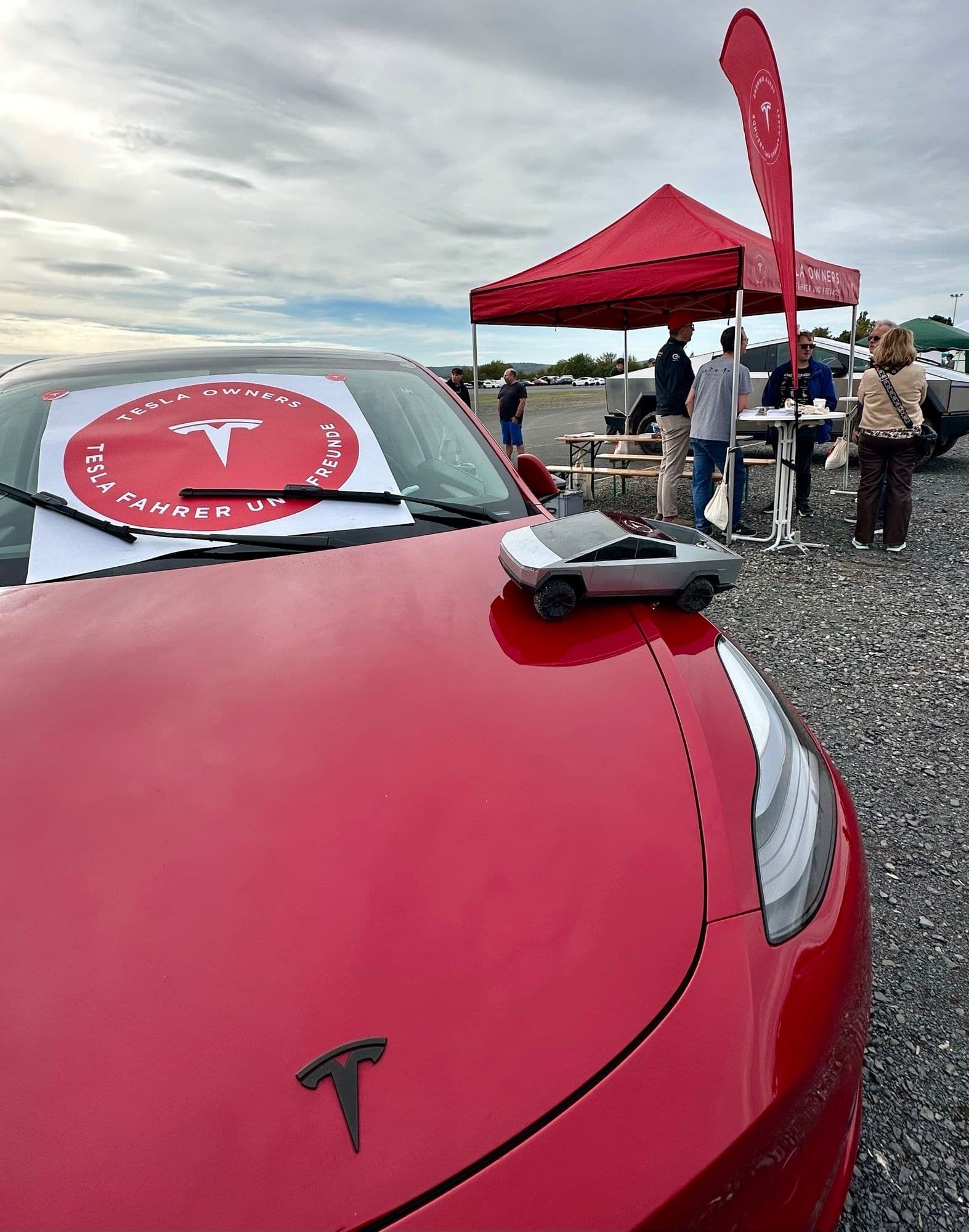 Roter Tesla mit Vereinsplakat hinter der Windschutzscheibe, auf der Motorhaube ein ferngesteuerter Cybertruck, dahinter der Vereinsstand