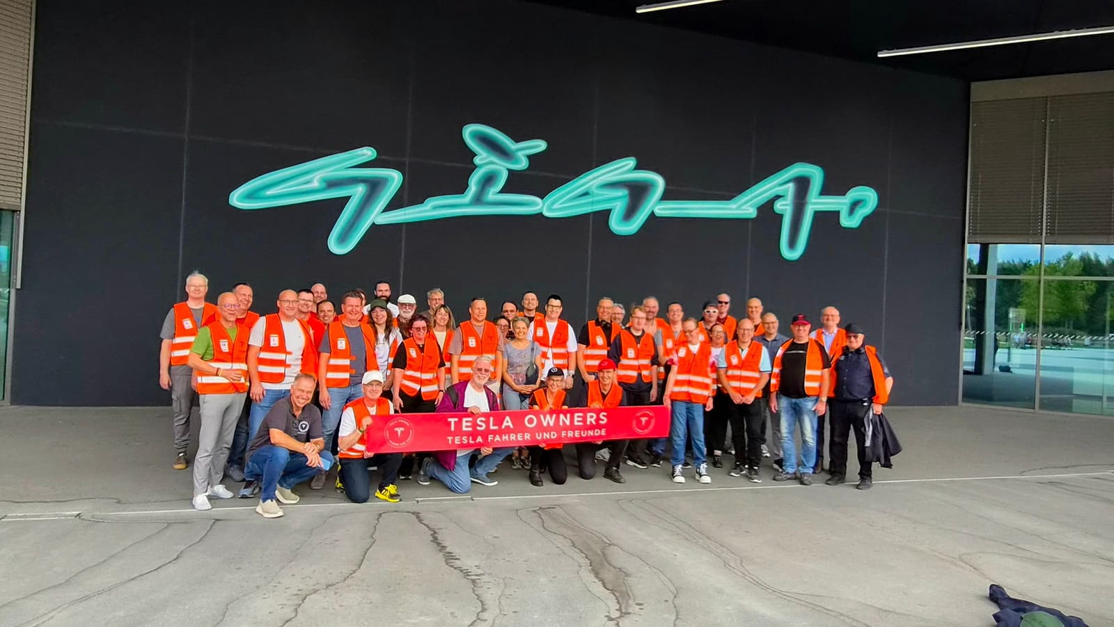 Die Gruppe in Warnwesten vor dem leuchtenden Giga-Schriftzug an der Fabrikwand, davor das Vereinsbanner