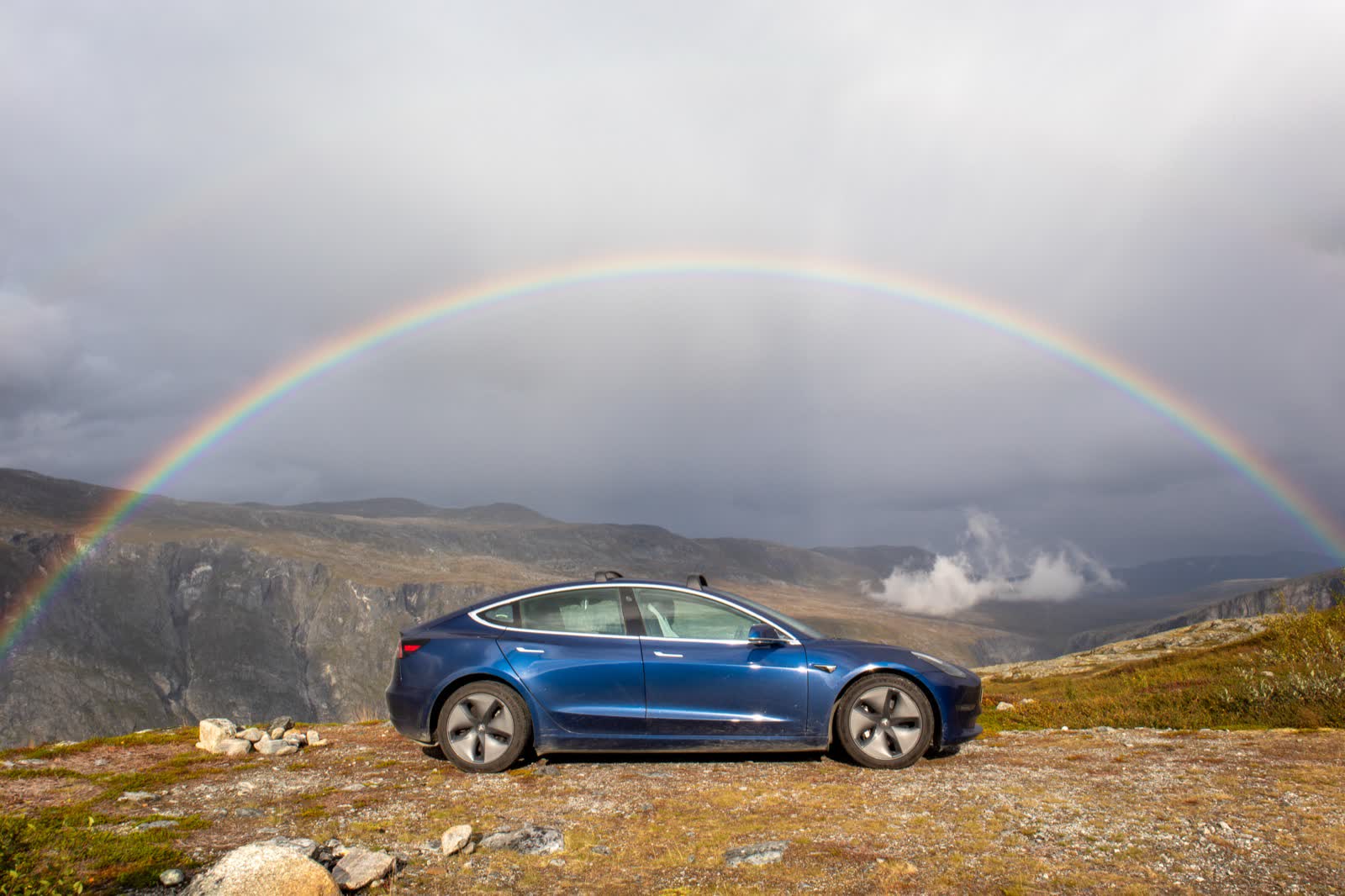 Blauer Tesla Model 3 auf einem Bergpass unter einem Regenbogen