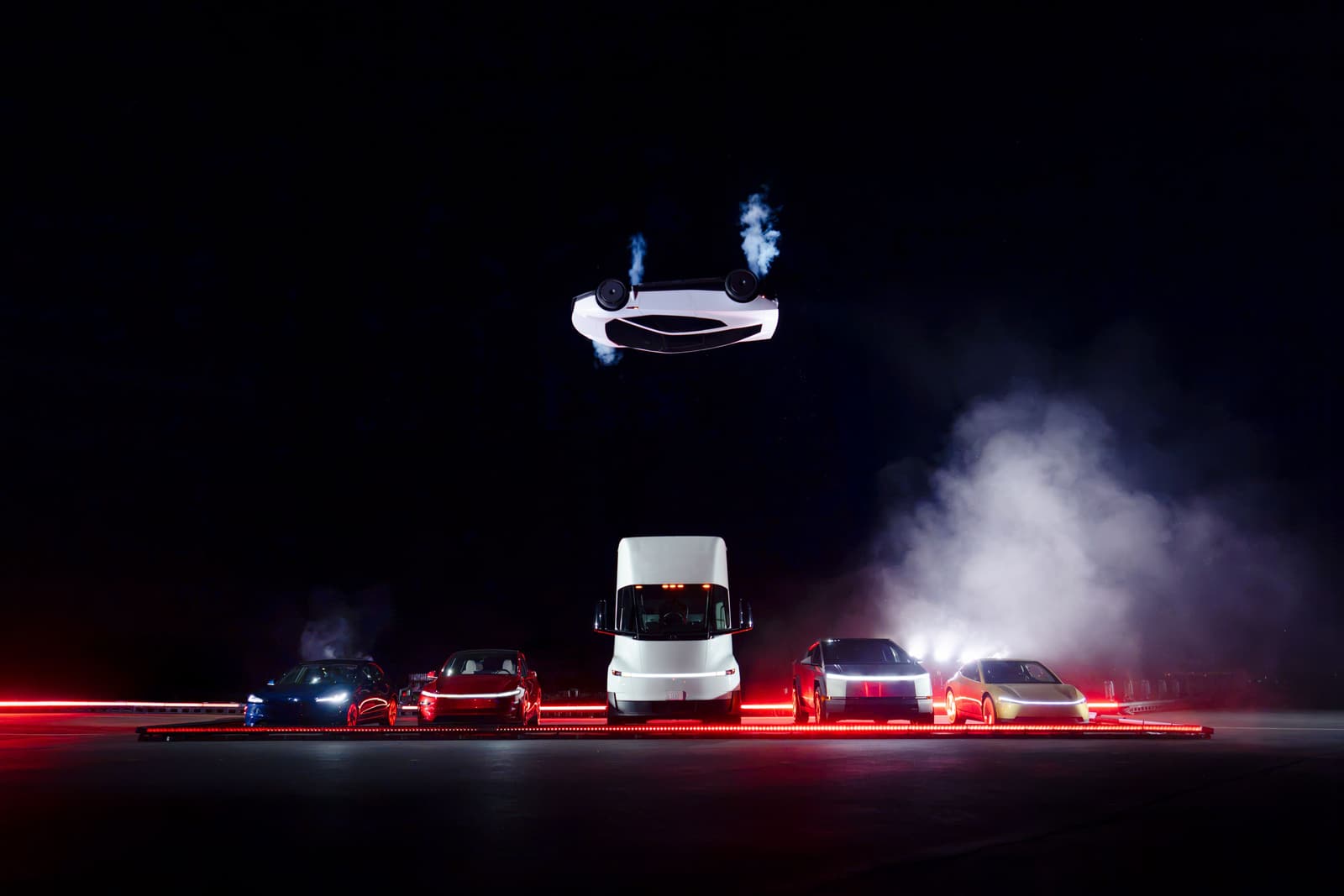 Der weiße Tesla Roadster kopfüber in der Luft über einer Reihe von Tesla-Fahrzeugen, darunter ein Semi, vor schwarzem Nachthimmel