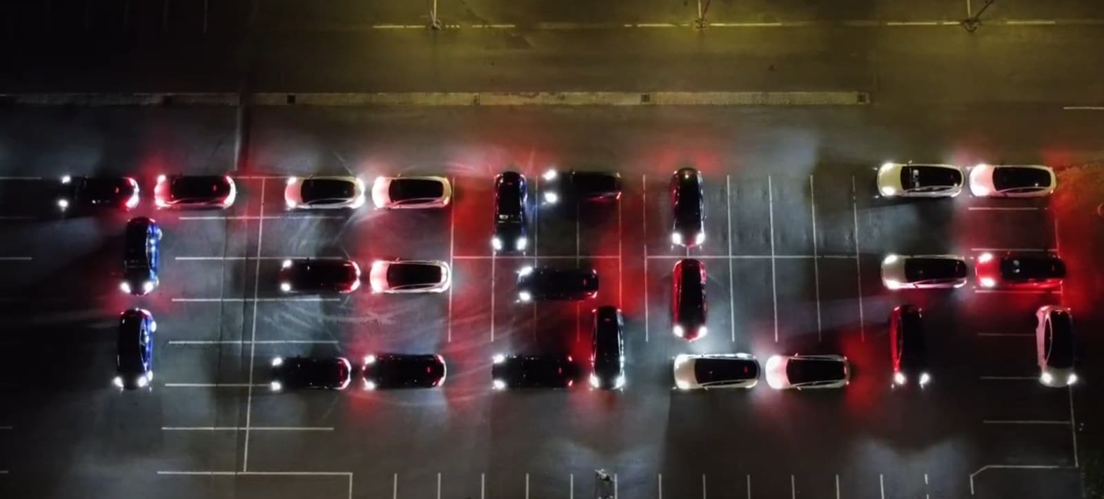 Luftaufnahme bei Nacht: rund dreißig Teslas stehen auf einem Parkplatz so, dass ihre Scheinwerfer Schriftzeichen bilden